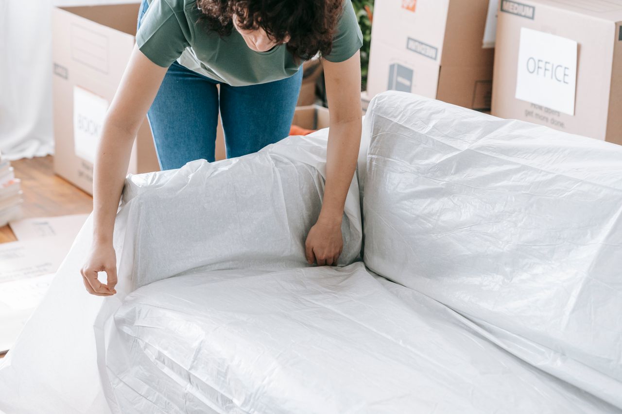 services-04 A woman carefully wraps a sofa in protective material while packing boxes in a bright room.